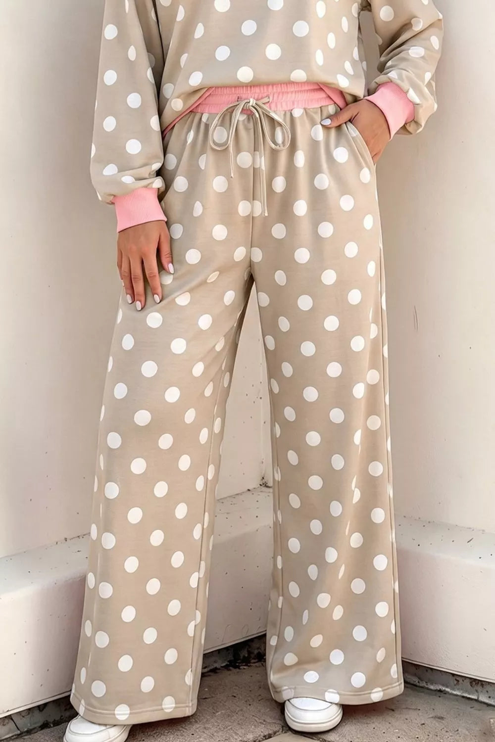 A person wearing beige wide-leg pajama pants with white polka dots and pink cuffs, paired with a matching long-sleeve top and white sneakers, standing against a light-colored wall.