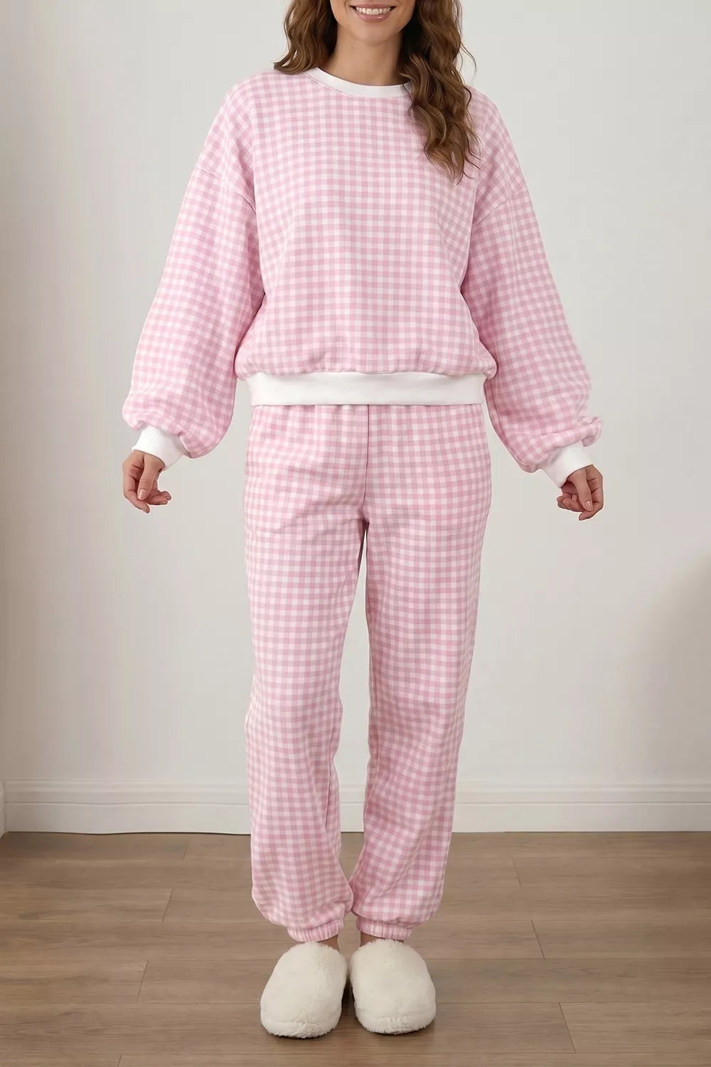 A woman modeling a pink and white gingham check pajama set, featuring a long-sleeve sweatshirt and matching pants, paired with white slippers in a simple indoor room setting.