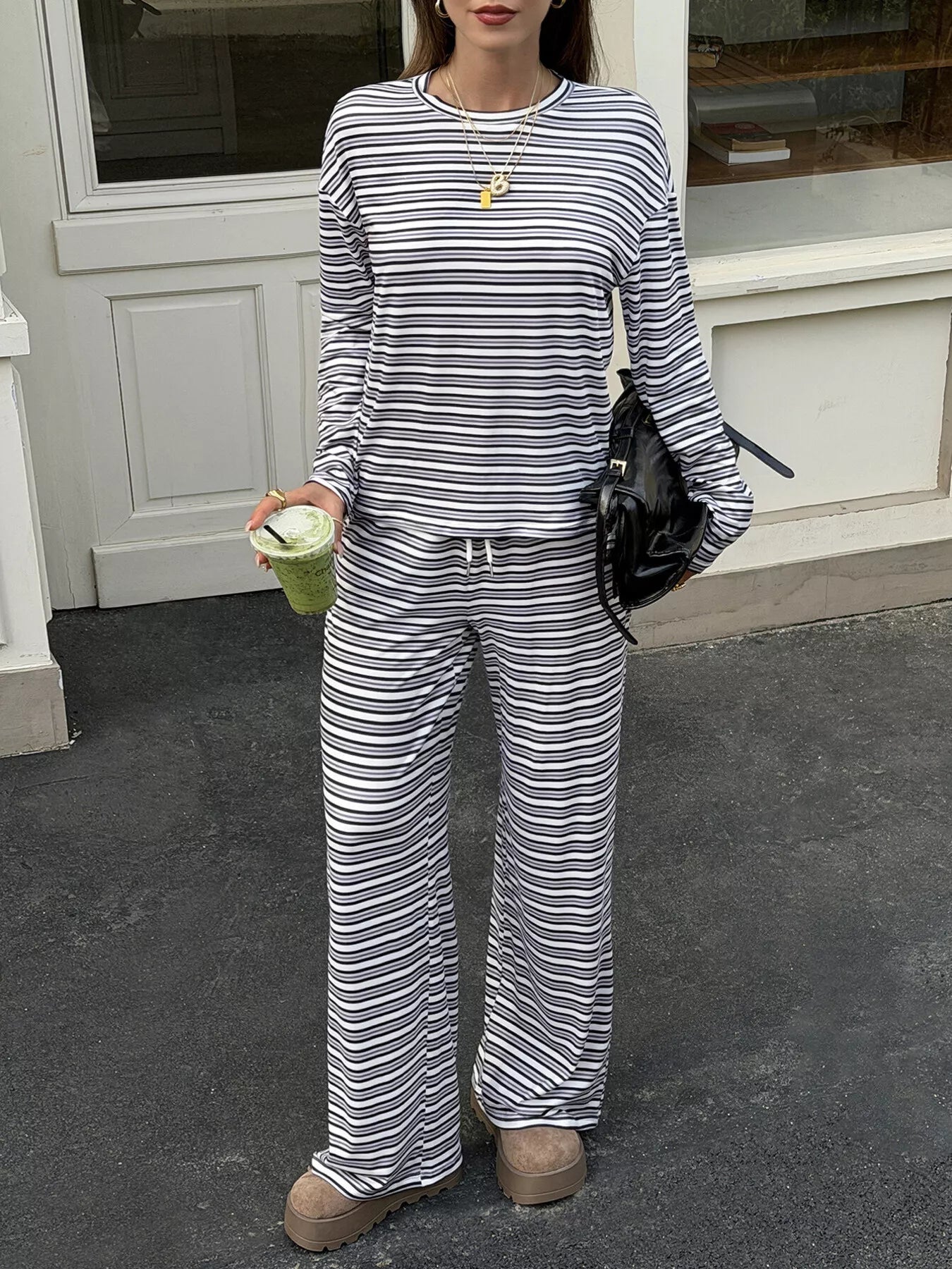 A woman wearing a matching long-sleeve striped lounge set with a crewneck top and wide-leg pants, holding a green drink and carrying a black shoulder bag, standing outdoors in front of a white building.