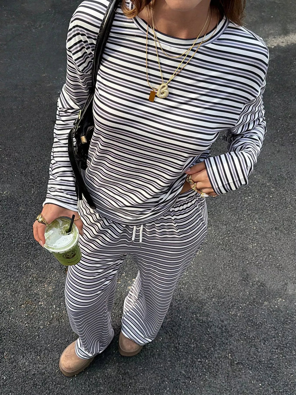 A person wearing a matching black and white striped long-sleeve top and drawstring sweatpants, standing outdoors on asphalt and holding a green iced coffee cup.