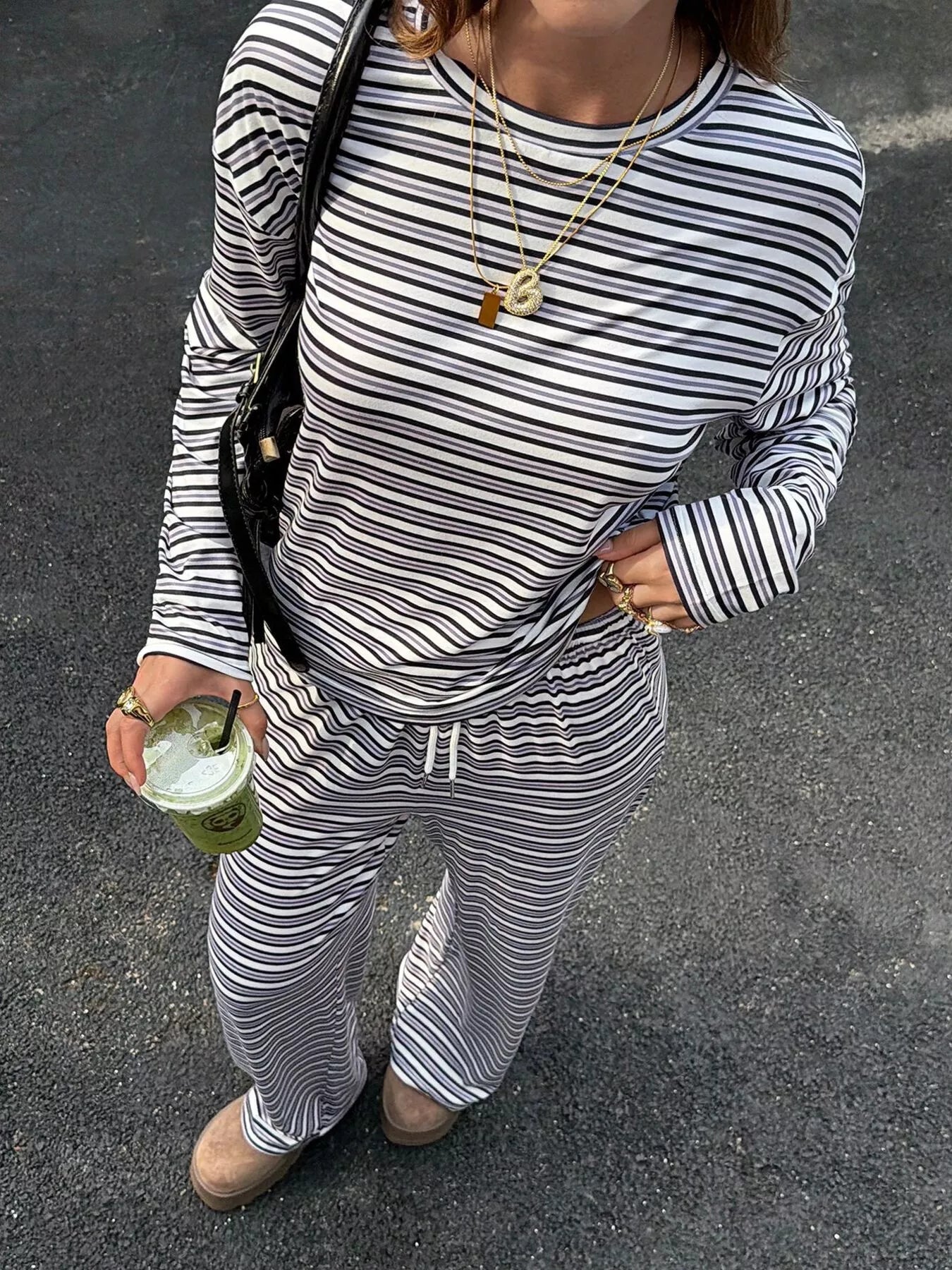 A person wearing a matching black and white striped long-sleeve top and drawstring sweatpants, standing outdoors on asphalt and holding a green iced coffee cup.