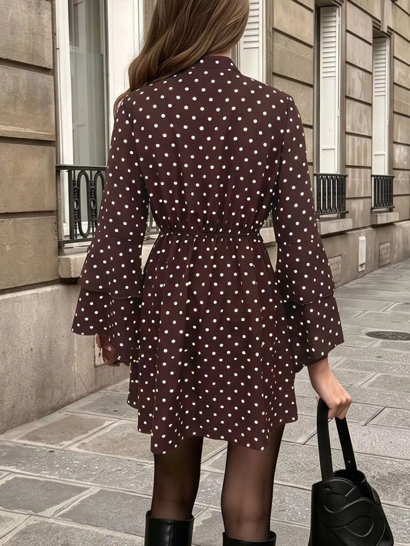A woman wearing a dark brown long-sleeve mini dress with white polka dots, shown from behind while standing on a city sidewalk next to a stone building.