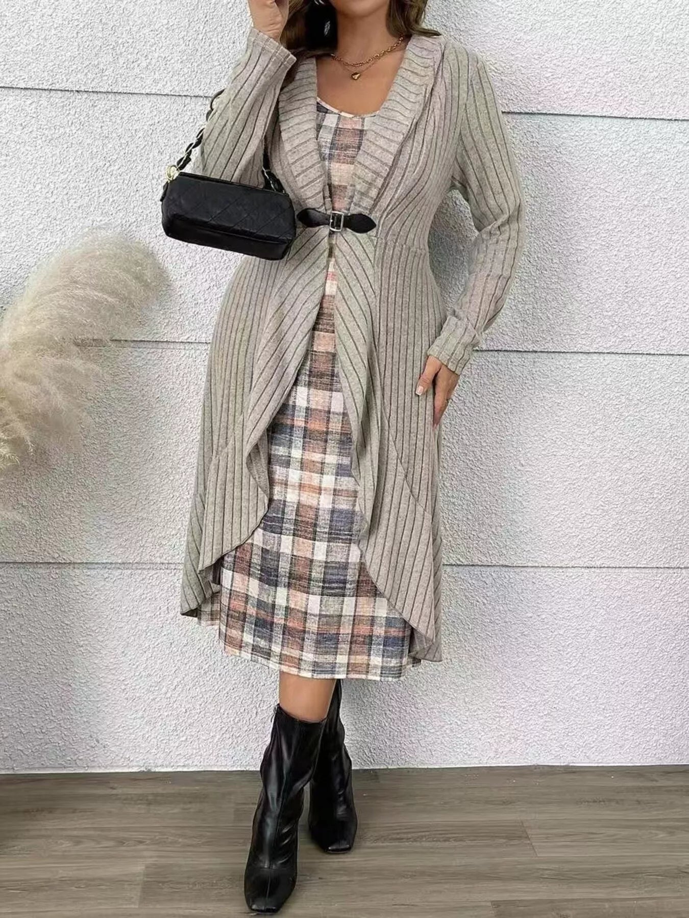 Woman wearing a long ribbed cardigan over a plaid dress with black boots against a light gray wall.