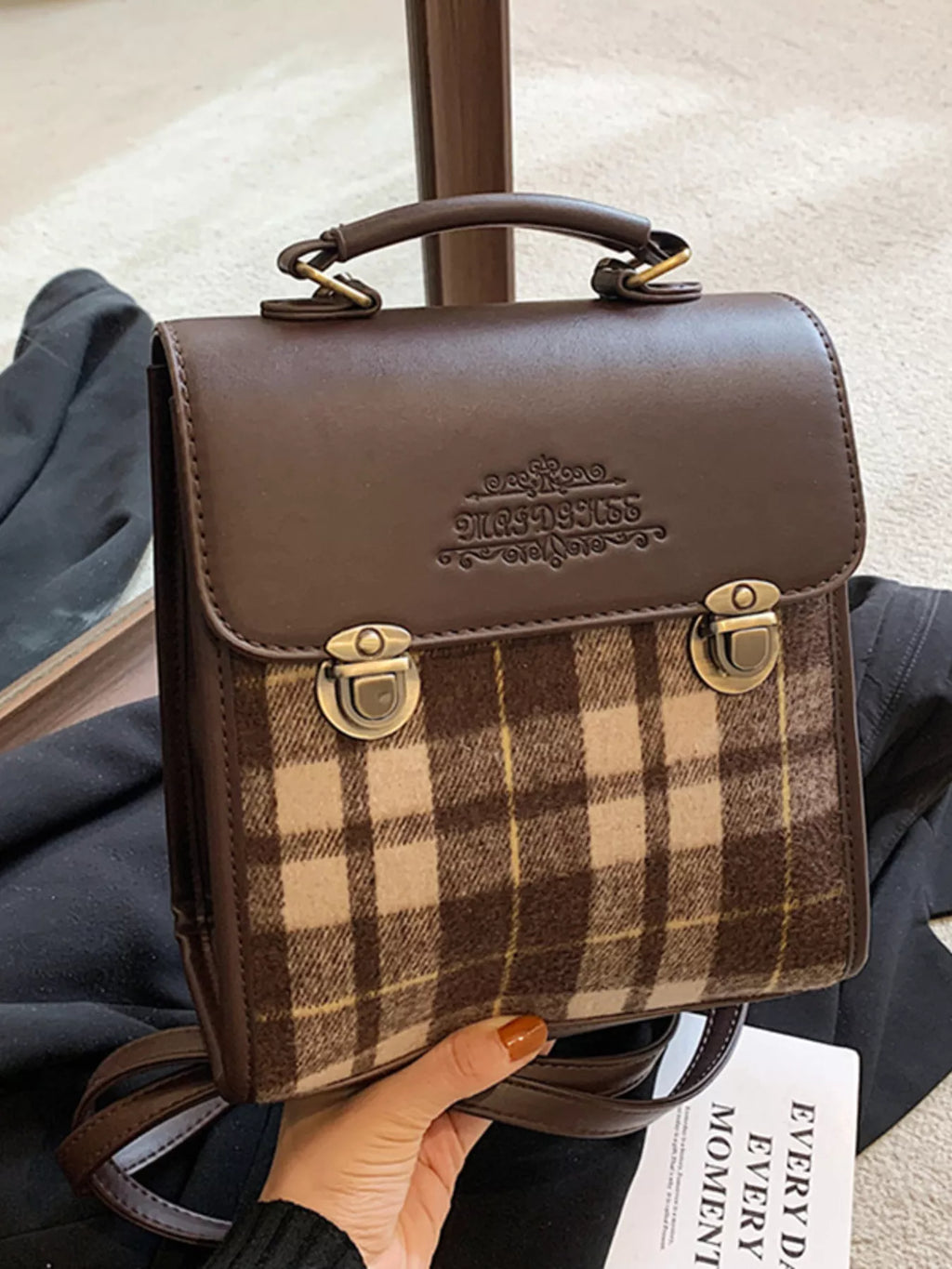 Brown leather and plaid fabric bag held by a person, with a visible brand logo.