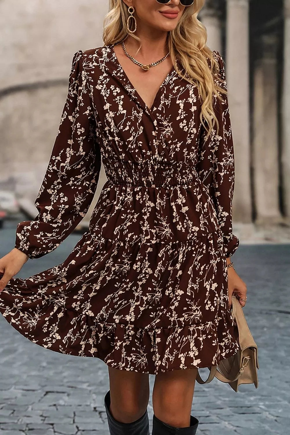 A woman wearing a long-sleeve brown wrap-style dress with a white floral print, standing outdoors on a cobblestone street.