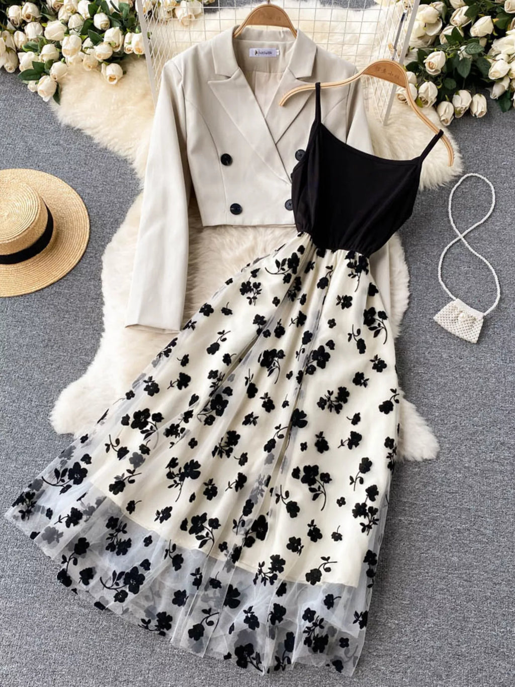 A styled outfit flat-lay featuring a cream double-breasted blazer, a black spaghetti-strap crop top, and a cream tulle midi skirt with a black floral print. The items are arranged on a gray textured rug with a white faux fur throw, a straw hat, and a small white purse.