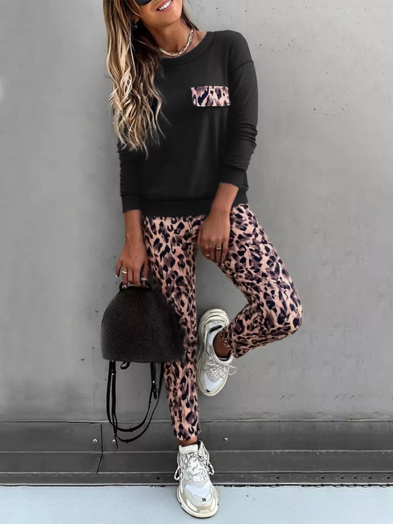 Person wearing a black sweatshirt with leopard print accents and leopard print pants, standing against a gray wall.