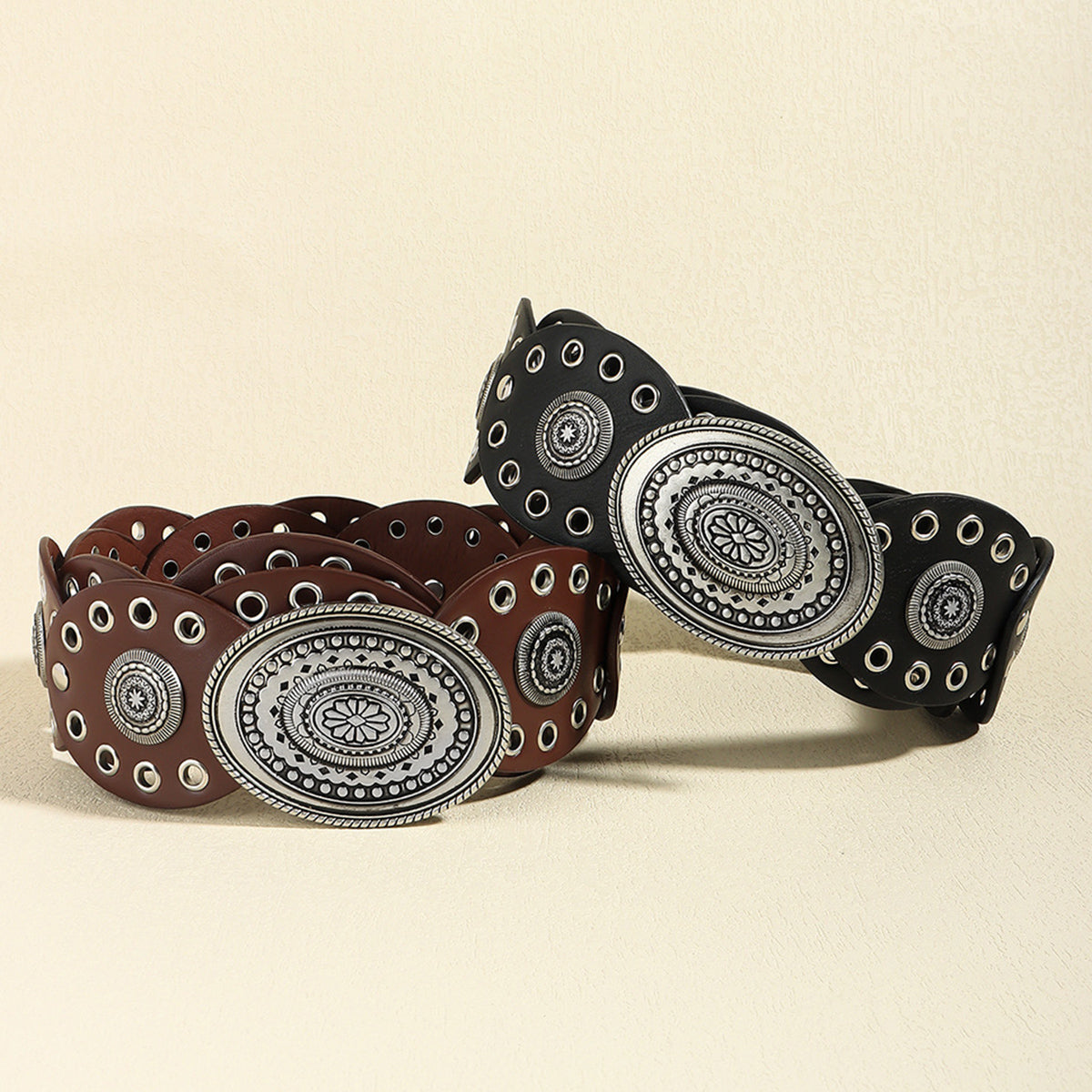 Two western-style leather belts with large oval silver-tone concho buckles and rows of silver grommets, one brown and one black, displayed on a light beige textured background.