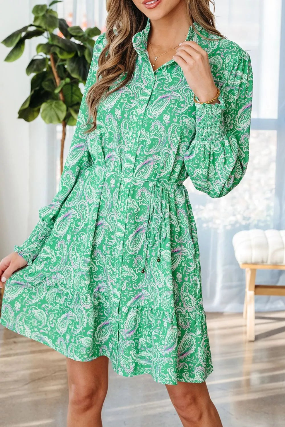 A model wearing a green long-sleeve mini dress with a white paisley pattern, standing in a bright indoor room with a plant and window in the background.