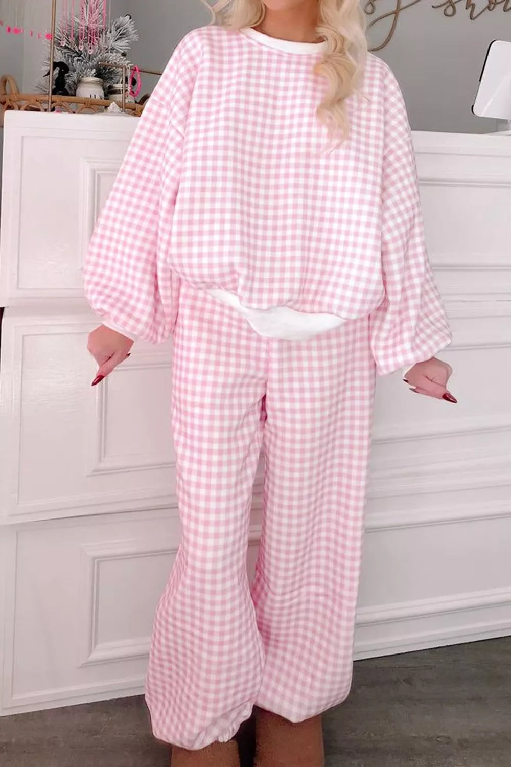 A woman wearing a pink and white gingham two-piece lounge set with a long-sleeve top and matching wide-leg pants, standing in front of a white paneled cabinet.