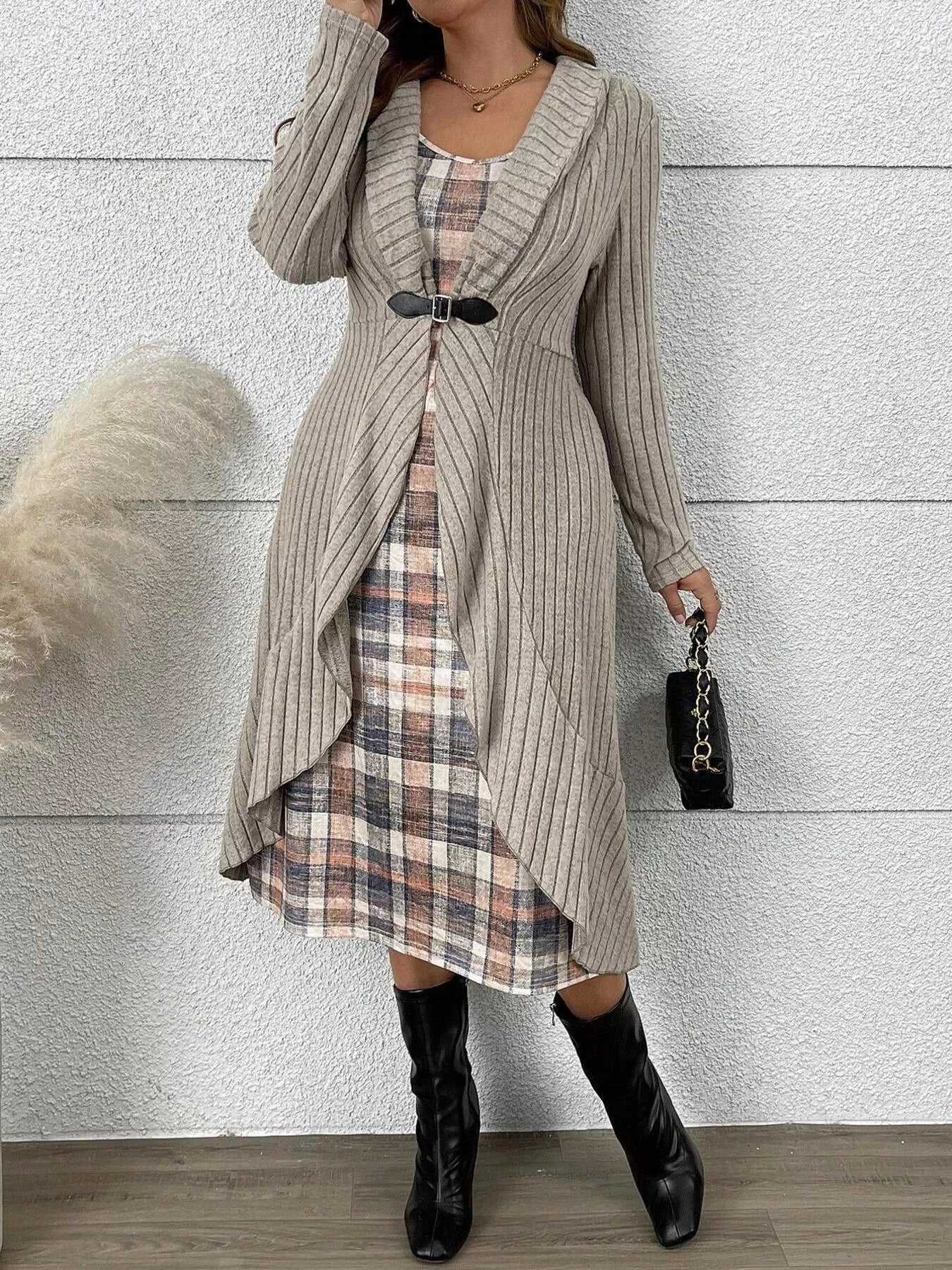 A woman models a long beige ribbed cardigan with vertical stripes over a plaid dress, paired with black knee-high boots and holding a small black handbag, set against a textured light gray wall.
