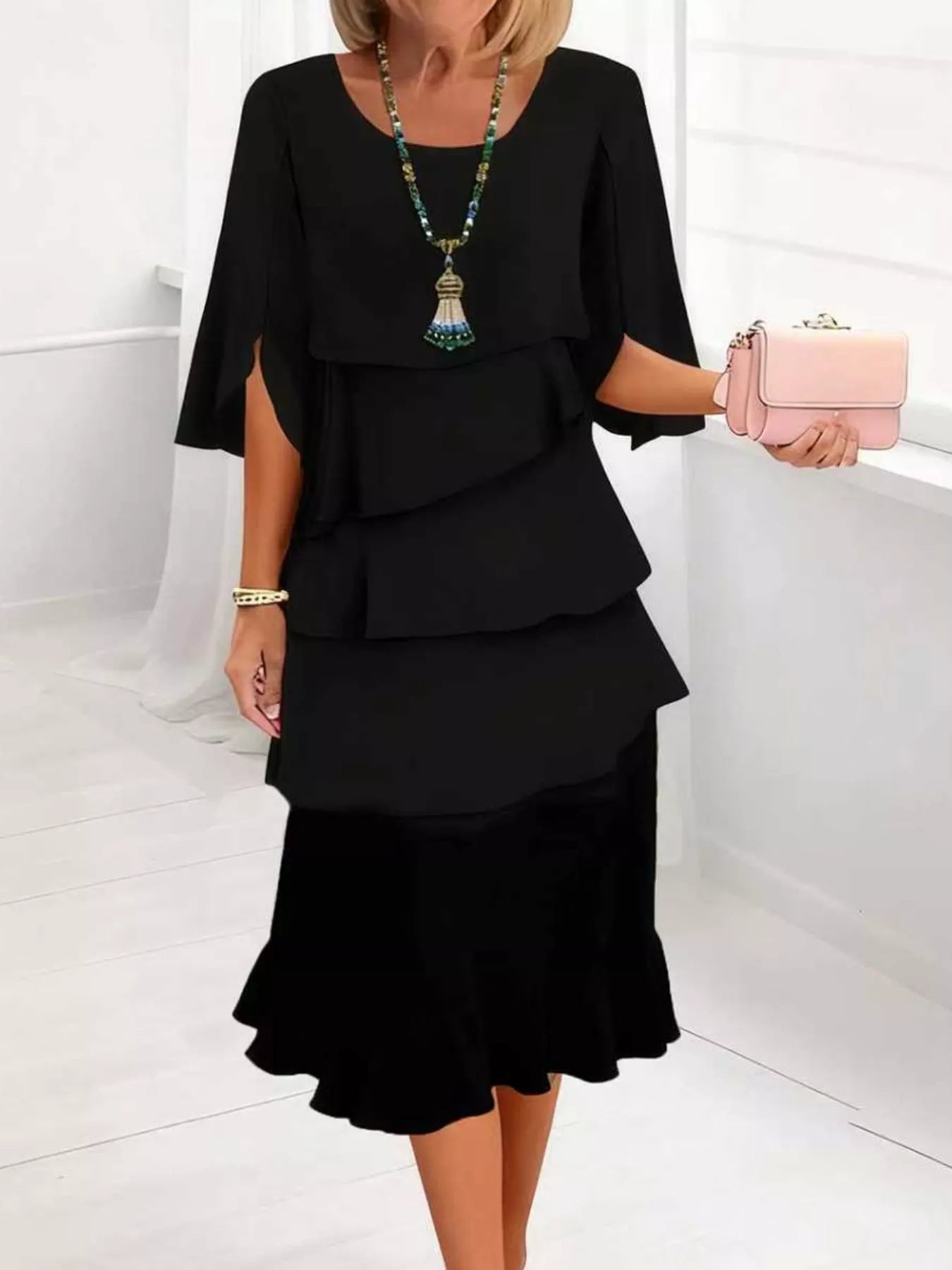A woman wearing a black tiered dress with wide sleeves and a ruffled hem, holding a small pink clutch bag in a bright white indoor setting.