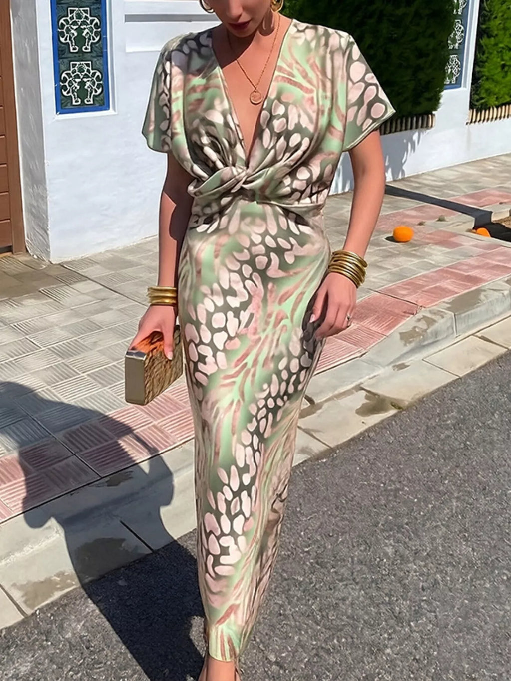A woman wearing a short-sleeve, V-neck maxi dress with a twist-front detail and an abstract spotted pattern in sage green, beige, and black, styled with gold jewelry and a metallic clutch on a sunny outdoor walkway.