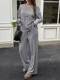 A woman wearing a matching long-sleeve striped lounge set with wide-leg pants, standing outdoors in front of a white building. She is holding a green iced coffee and carrying a black handbag.