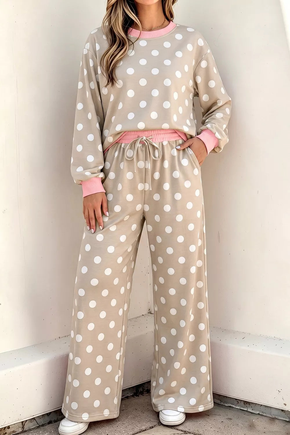 A woman wearing a beige and white polka dot lounge set consisting of a long-sleeve sweatshirt and matching wide-leg pants, with pink trim and a pink drawstring waist, standing against a light-colored wall.