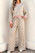 A woman wearing a beige and white polka dot lounge set consisting of a long-sleeve sweatshirt and matching wide-leg pants, with pink trim and a pink drawstring waist, standing against a light-colored wall.