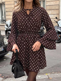 A woman wearing a dark brown long-sleeve mini dress with white polka dots, featuring a keyhole tie neckline and flared bell sleeves. She is holding a black handbag on a city street with parked scooters and a building in the background.