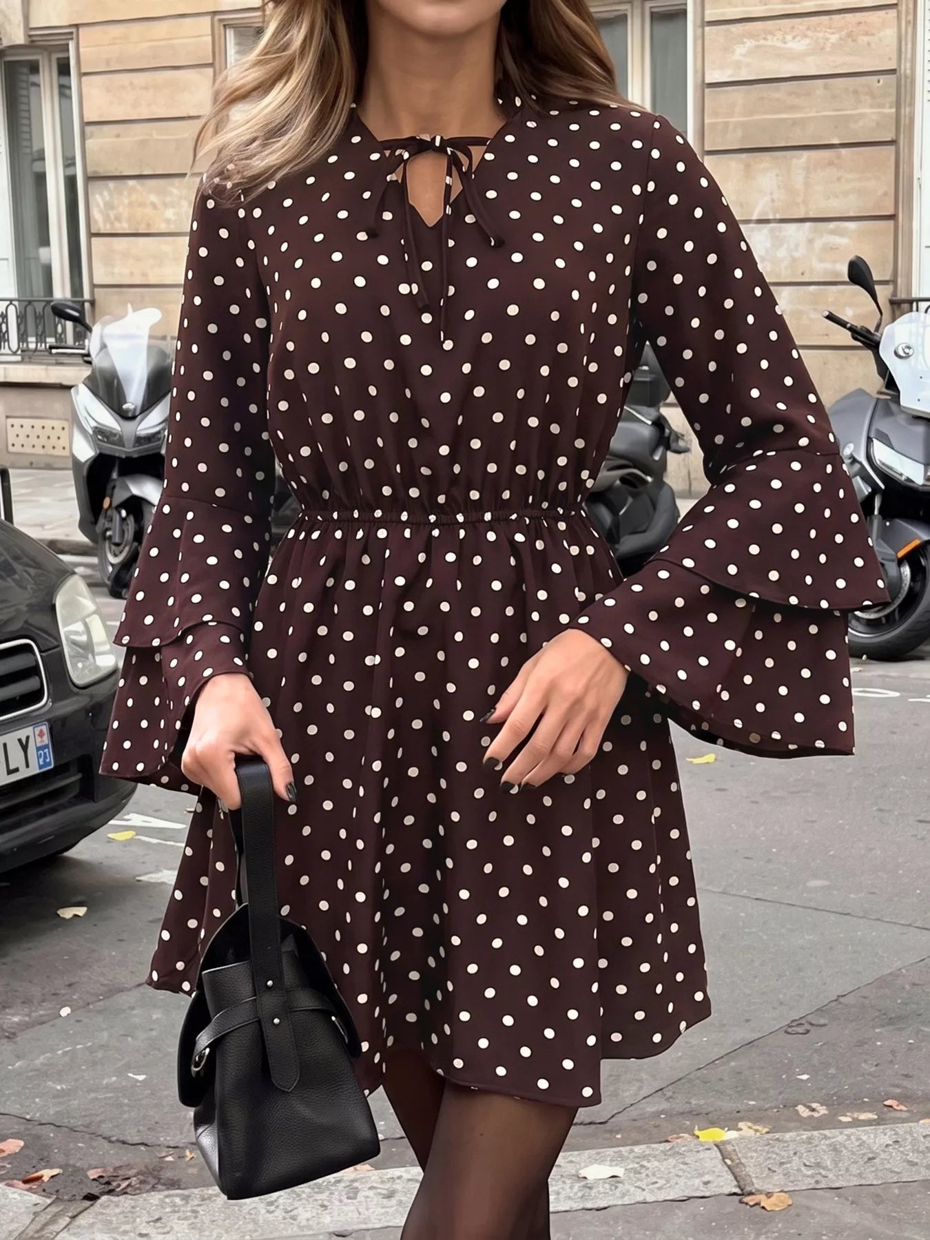 A woman wearing a dark brown long-sleeve mini dress with white polka dots, featuring a keyhole tie neckline and flared bell sleeves. She is holding a black handbag on a city street with parked scooters and a building in the background.