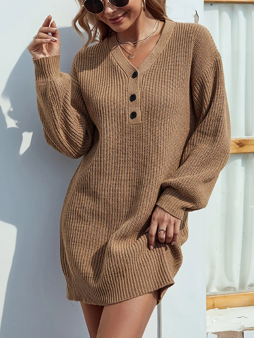 Woman wearing a brown knit dress with black buttons against a white wall.