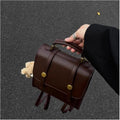 A person holds a dark brown faux-leather top-handle backpack with gold-tone snap buttons and straps, photographed outdoors on a gray asphalt surface.