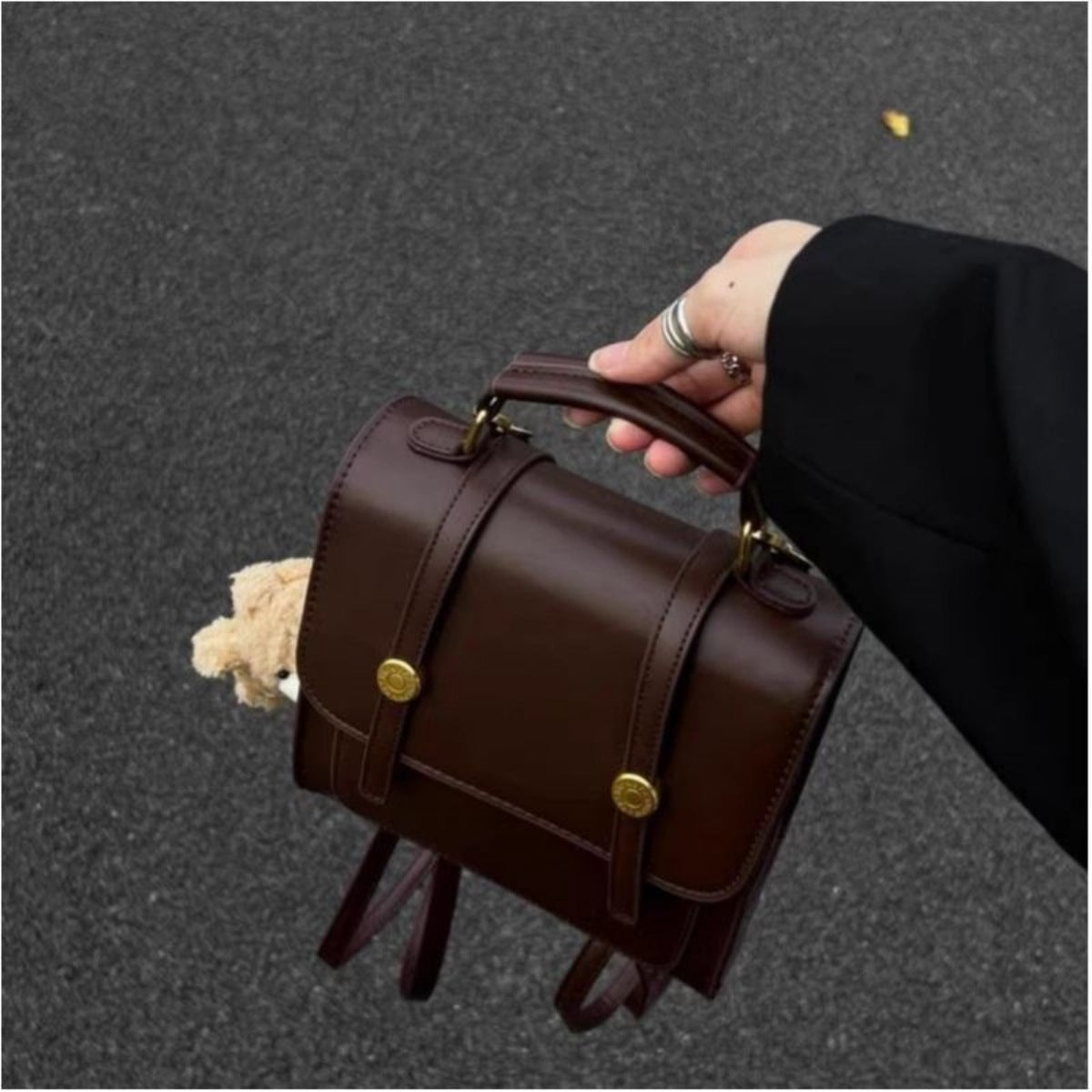 A person holds a dark brown faux-leather top-handle backpack with gold-tone snap buttons and straps, photographed outdoors on a gray asphalt surface.