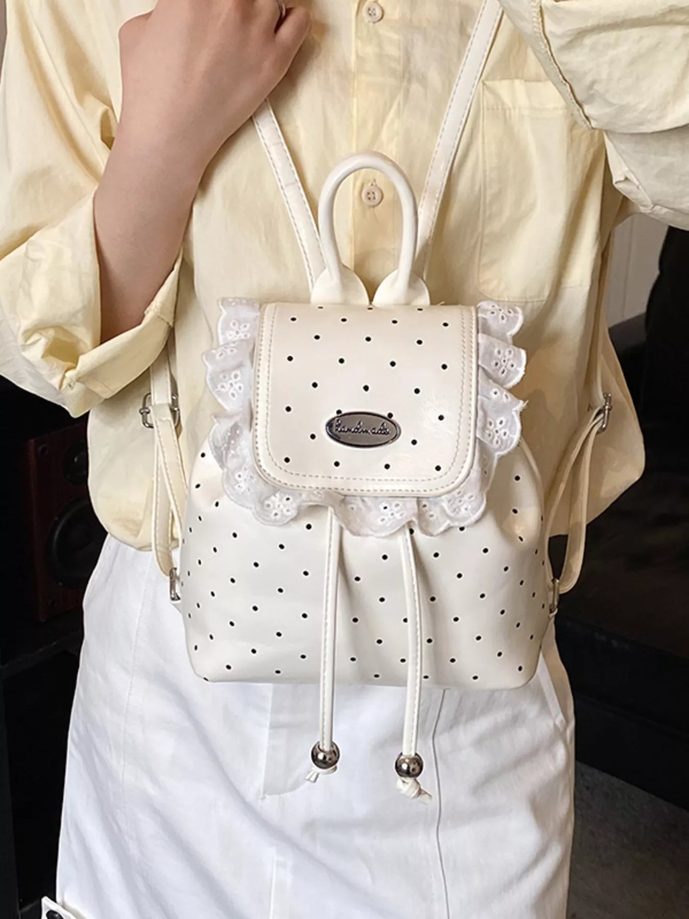 White polka dot backpack with lace details held by a model wearing a yellow shirt.