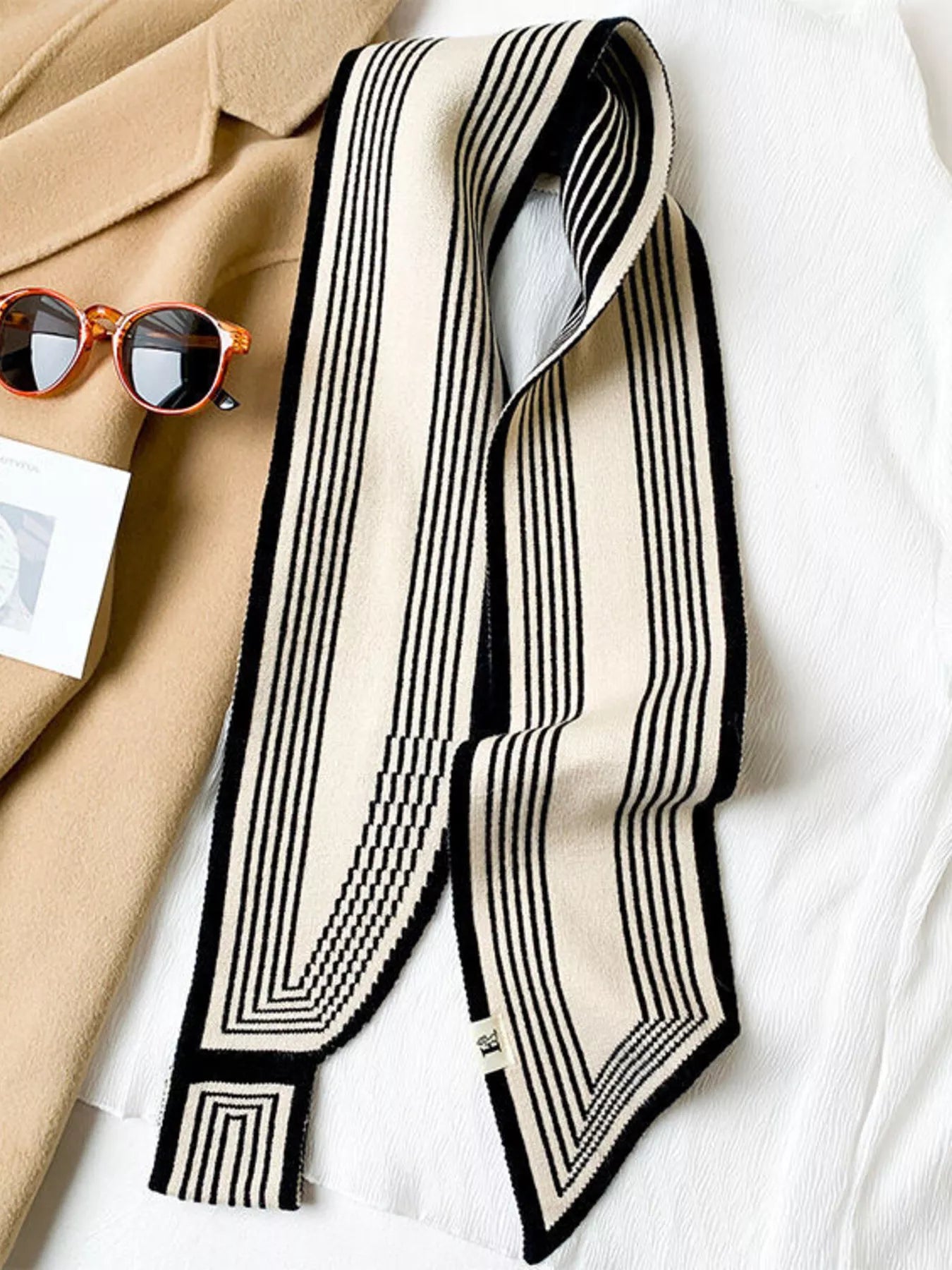Black and white striped scarf on a white surface with sunglasses and a beige coat.