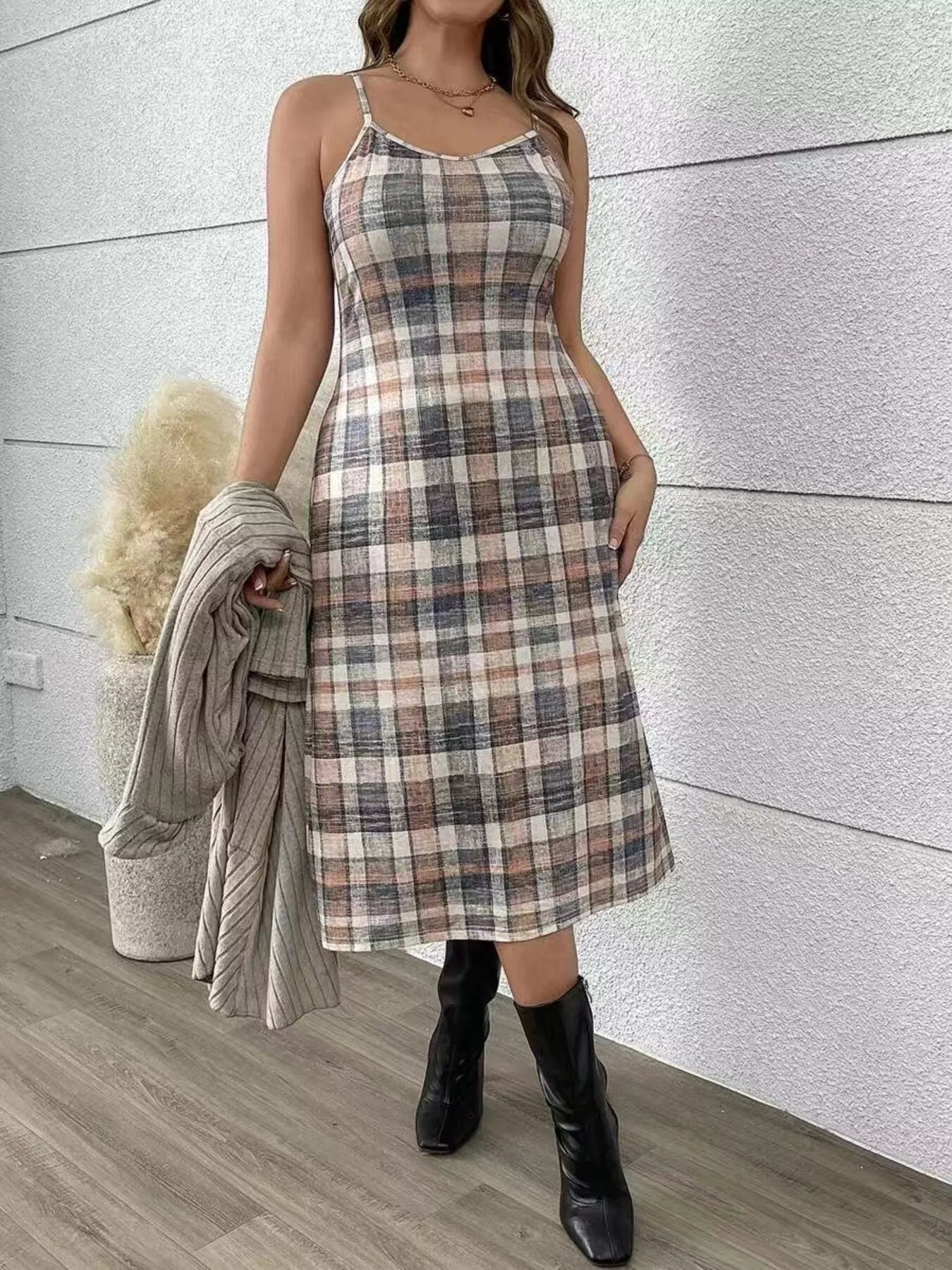 Woman wearing a plaid dress with a gray sweater over her shoulder against a light-colored wall.
