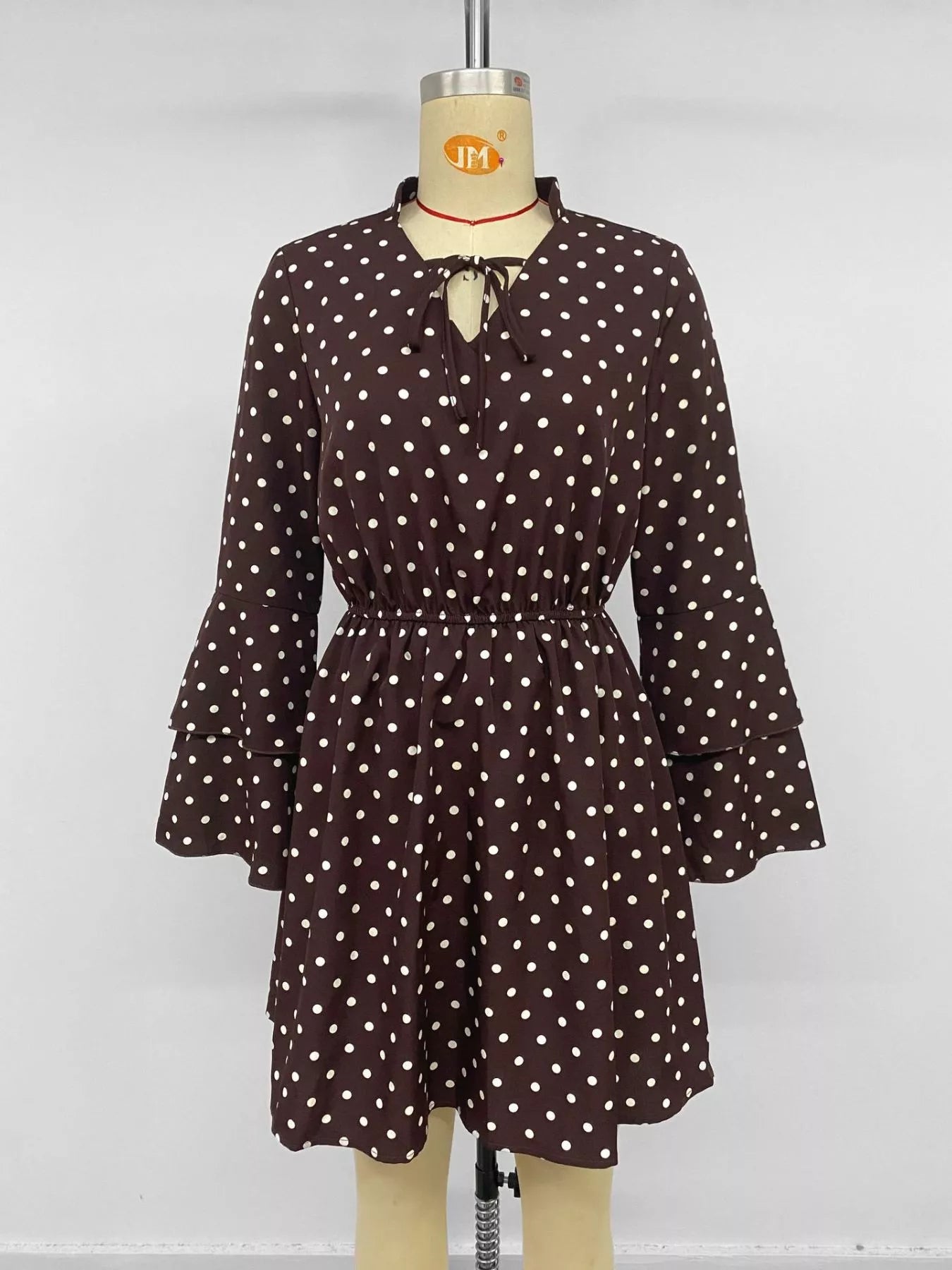 A brown long-sleeve mini dress with white polka dots, featuring a tie-neck detail and flared bell sleeves, displayed on a mannequin.