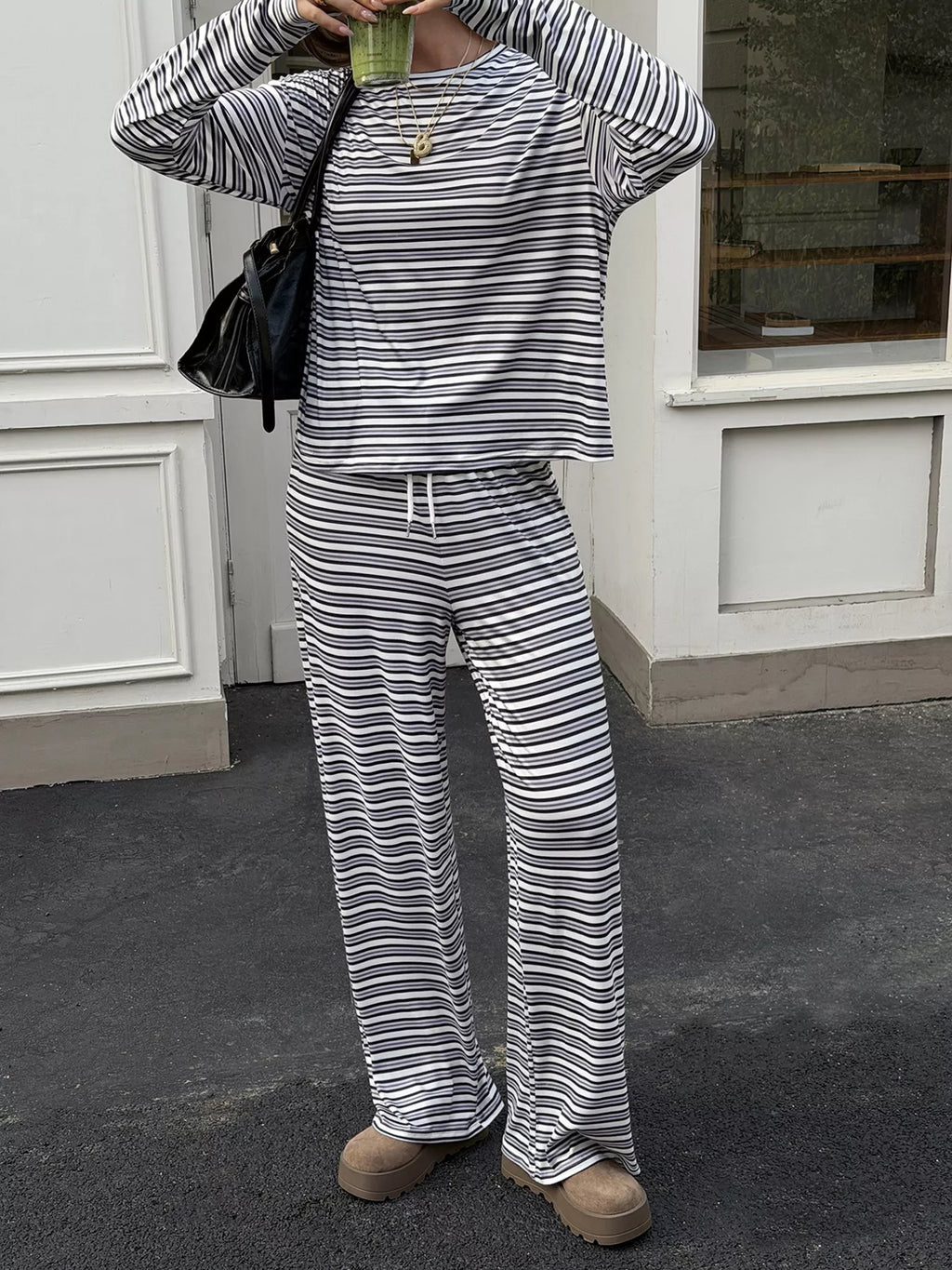A person wearing a matching striped lounge set with a long-sleeve top and wide-leg pants, standing outdoors in front of a white building. The outfit has thin horizontal black and off-white stripes, and the person is also holding a green drink and wearing a black shoulder bag.