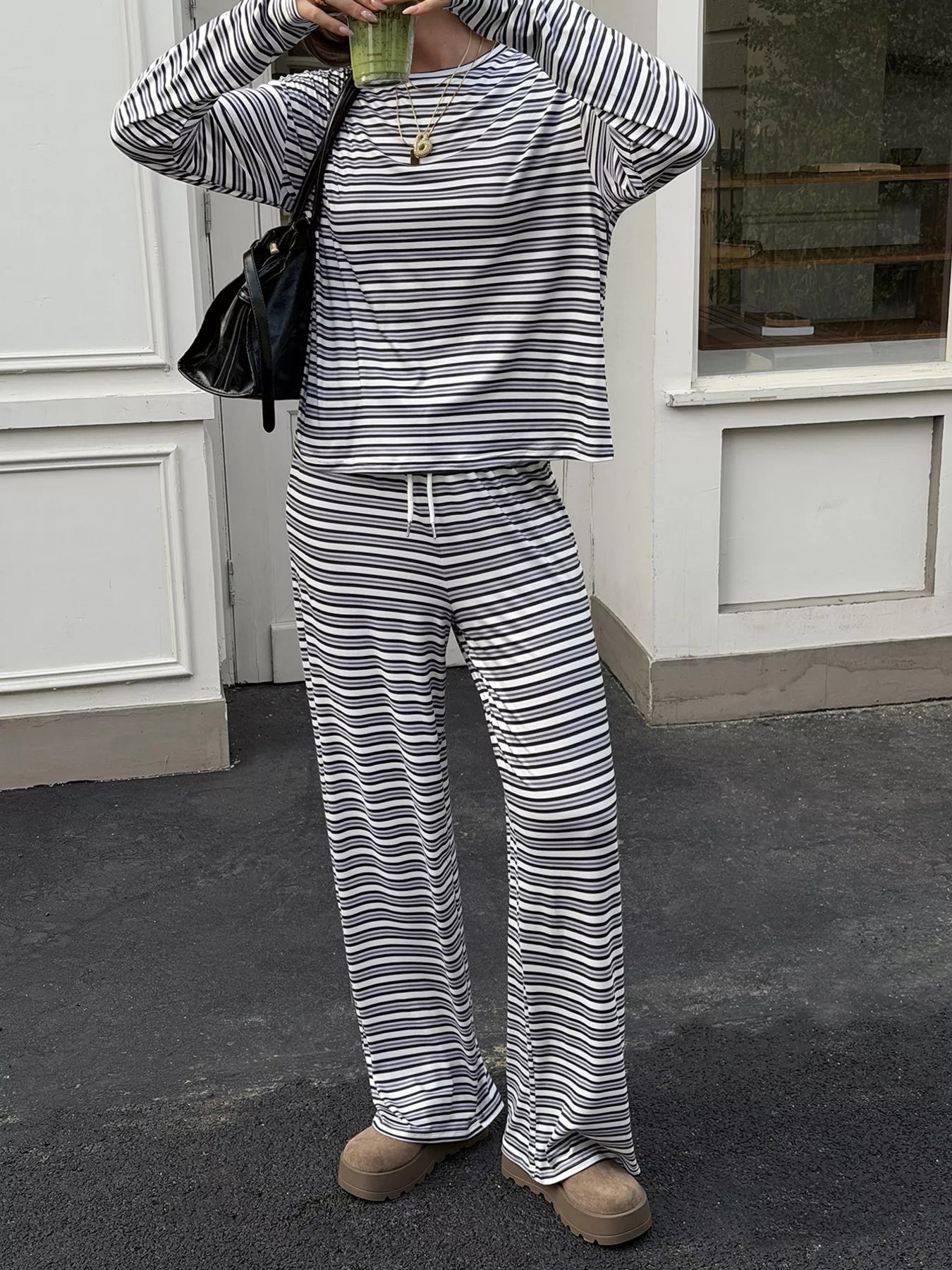 A person wearing a matching striped lounge set with a long-sleeve top and wide-leg pants, standing outdoors in front of a white building. The outfit has thin horizontal black and off-white stripes, and the person is also holding a green drink and wearing a black shoulder bag.