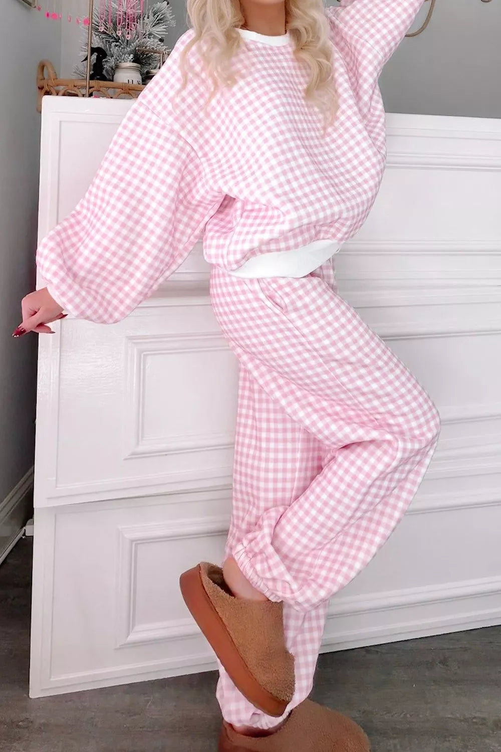 A person wearing a pink and white gingham matching lounge set with a cropped sweatshirt and wide-leg pants, styled with a white belt and tan slippers, standing in front of a white paneled cabinet.