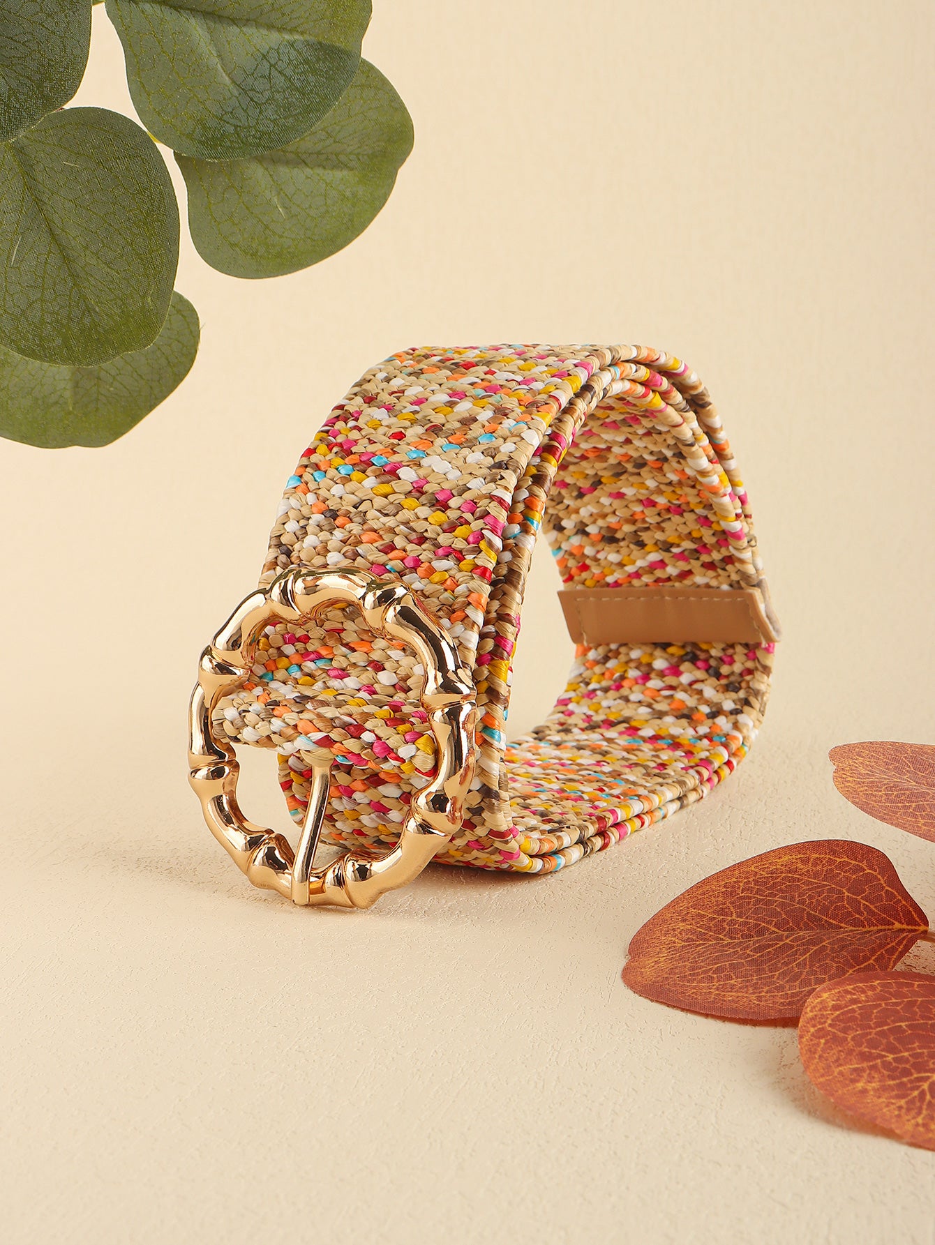 Multicolored woven belt with a gold buckle on a beige background with leaves and wood pieces.