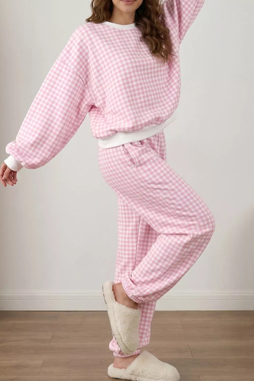 A woman modeling a pink and white gingham two-piece pajama set with a sweatshirt and matching pants, paired with cream slippers, in a minimalist indoor setting.