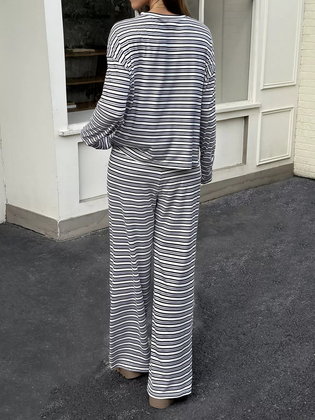 A woman viewed from behind wearing a matching two-piece loungewear set with horizontal navy and off-white stripes, consisting of a long-sleeve top and wide-leg pants, standing on a dark pavement in front of a white building.