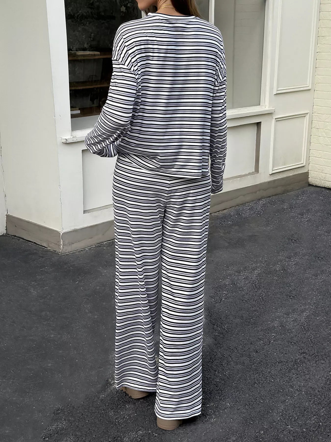 A woman viewed from behind wearing a matching two-piece loungewear set with horizontal navy and off-white stripes, consisting of a long-sleeve top and wide-leg pants, standing on a dark pavement in front of a white building.